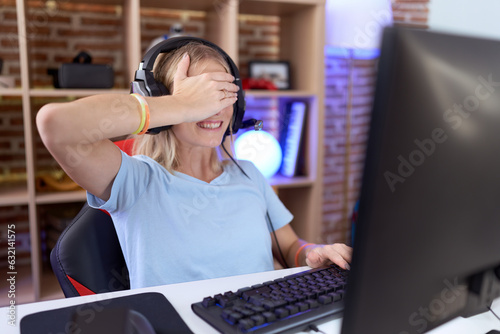 Young caucasian woman playing video games wearing headphones smiling and laughing with hand on face covering eyes for surprise. blind concept.