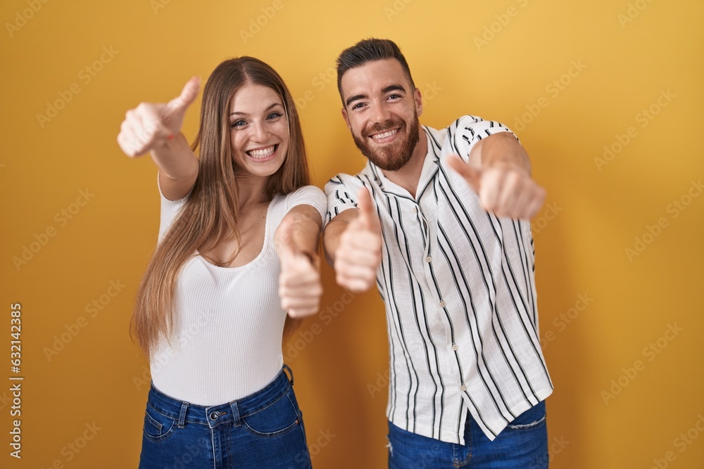 Young couple standing over yellow background approving doing positive gesture with hand, thumbs up smiling and happy for success. winner gesture. Young couple standing over yellow background approving doing positive gesture with hand, thumbs up smiling and happy for success. winner gesture.
