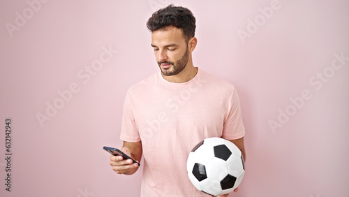 Young hispanic man using smartphone holding soccer ball over isolated pink background © Krakenimages.com