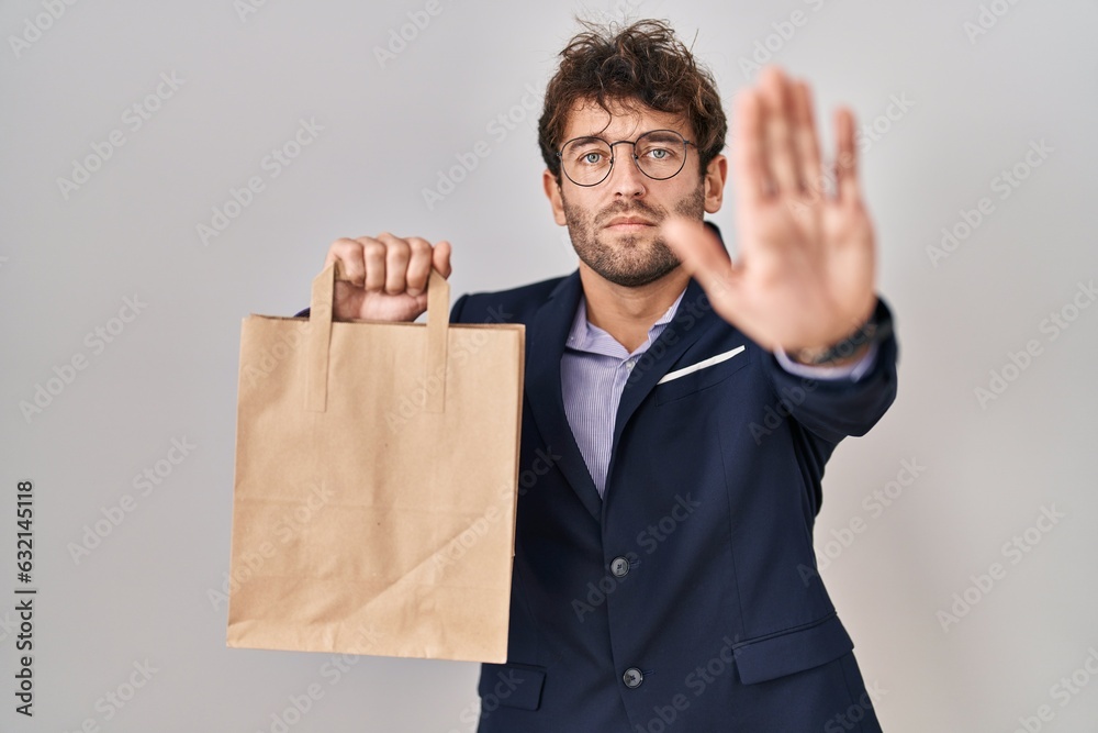 Hispanic business man holding delivery bag with open hand doing stop sign with serious and confident expression, defense gesture Hispanic business man holding delivery bag with open hand doing stop sign with serious and confident expression, defense gesture