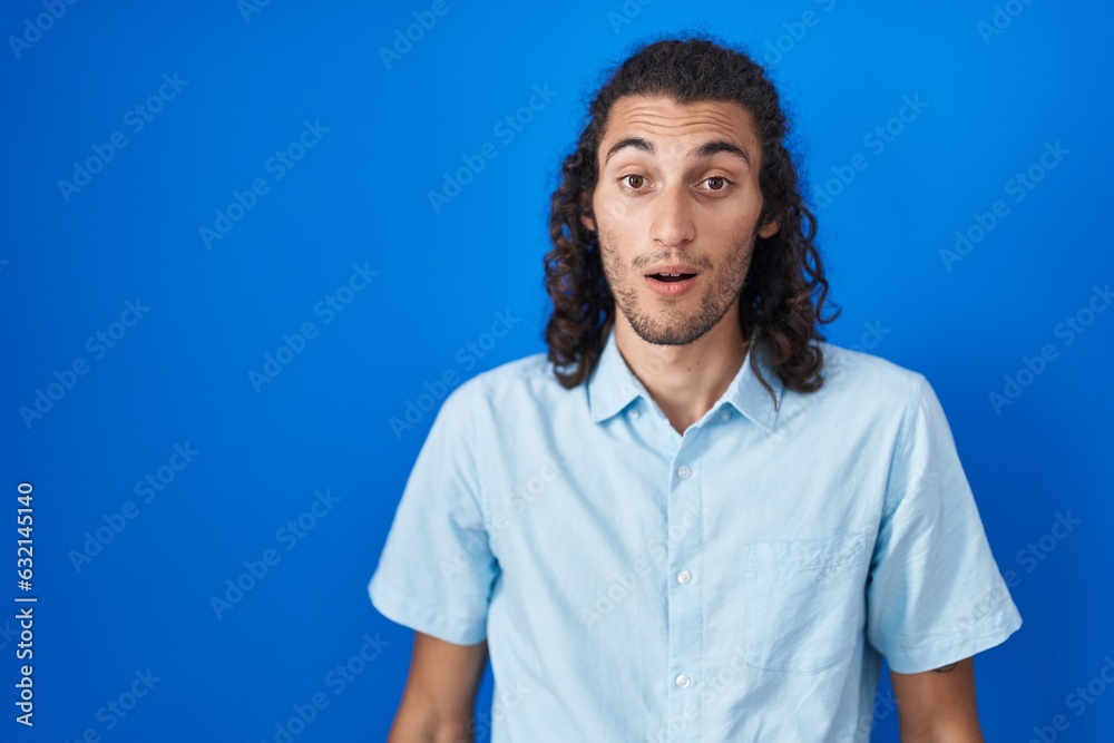 Young hispanic man standing over blue background afraid and shocked with surprise and amazed expression, fear and excited face. Young hispanic man standing over blue background afraid and shocked with surprise and amazed expression, fear and excited face.