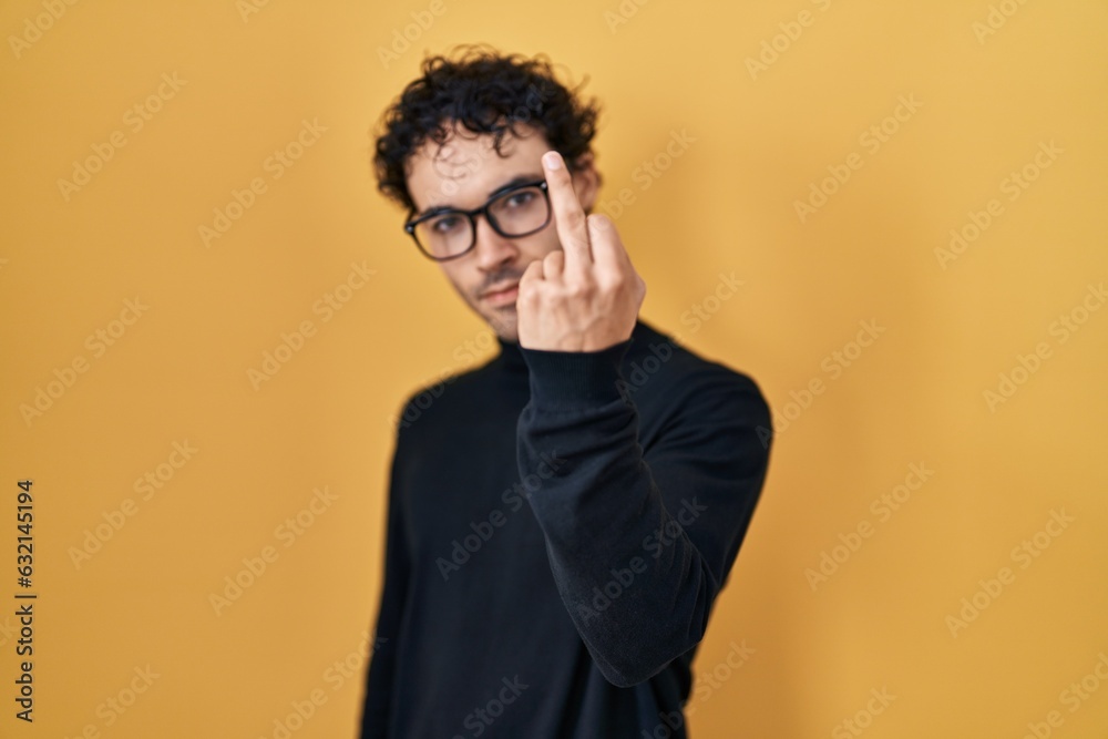 Hispanic man standing over yellow background showing middle finger, impolite and rude fuck off expression