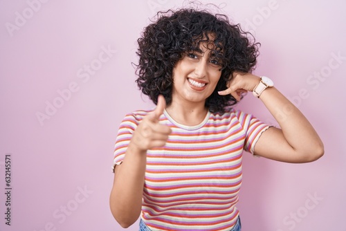 Young middle east woman standing over pink background smiling doing talking on the telephone gesture and pointing to you. call me.