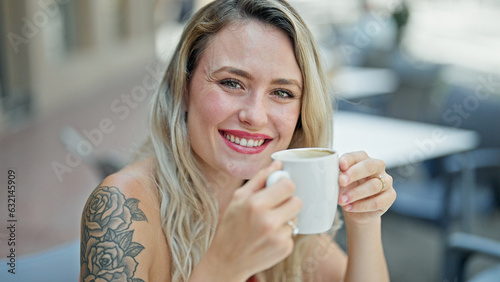 Young blonde woman drinking coffee sitting on table smiling at coffee shop terrace