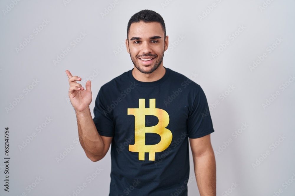 Young hispanic man wearing bitcoin t shirt cheerful with a smile on face pointing with hand and finger up to the side with happy and natural expression Young hispanic man wearing bitcoin t shirt cheerful with a smile on face pointing with hand and finger up to the side with happy and natural expression