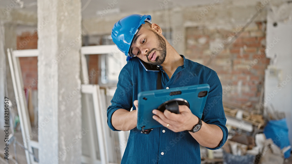 Fototapeta premium Young hispanic man architect using touchpad device speaking on the phone at construction site