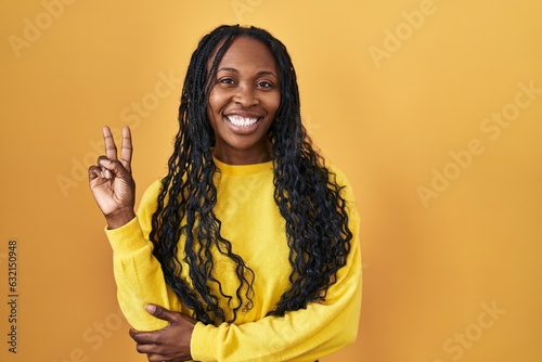 African woman standing over yellow background smiling with happy face winking at the camera doing victory sign. number two. © Krakenimages.com