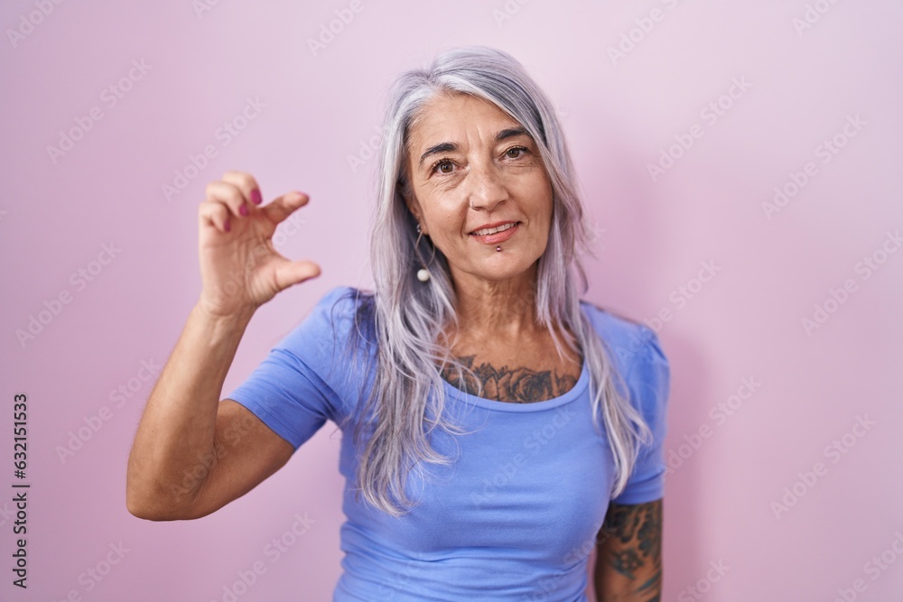 Middle age woman with tattoos standing over pink background smiling and confident gesturing with hand doing small size sign with fingers looking and the camera. measure concept. Middle age woman with tattoos standing over pink background smiling and confident gesturing with hand doing small size sign with fingers looking and the camera. measure concept.