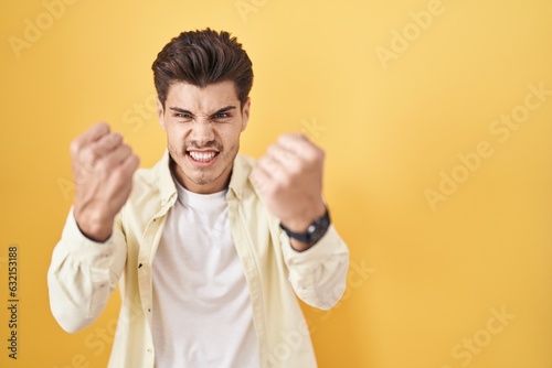 Young hispanic man standing over yellow background angry and mad raising fists frustrated and furious while shouting with anger. rage and aggressive concept.