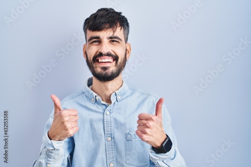 Young hispanic man with beard standing over blue background success sign doing positive gesture with hand, thumbs up smiling and happy. cheerful expression and winner gesture.