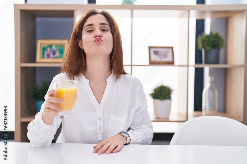 Brunette woman drinking glass of orange juice looking at the camera blowing a kiss on air being lovely and sexy. love expression. Brunette woman drinking glass of orange juice looking at the camera blowing a kiss on air being lovely and sexy. love expression.