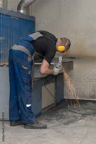 A male worker in uniform cutting a metal part with a cutter at the workplace at the factory. Sparks, flames and smoke from cutting