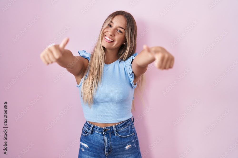 Young blonde woman standing over pink background approving doing positive gesture with hand, thumbs up smiling and happy for success. winner gesture. Young blonde woman standing over pink background approving doing positive gesture with hand, thumbs up smiling and happy for success. winner gesture.