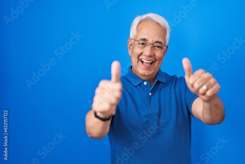 Middle age man with grey hair standing over blue background approving doing positive gesture with hand, thumbs up smiling and happy for success. winner gesture.