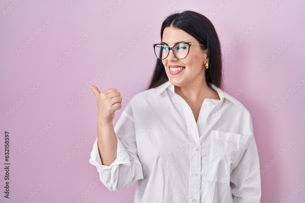 Young brunette woman standing over pink background smiling with happy face looking and pointing to the side with thumb up. Young brunette woman standing over pink background smiling with happy face looking and pointing to the side with thumb up.