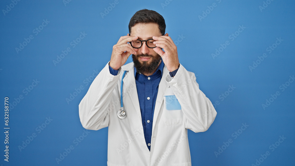 Young hispanic man doctor standing with serious expression wearing glasses over isolated blue background Young hispanic man doctor standing with serious expression wearing glasses over isolated blue background