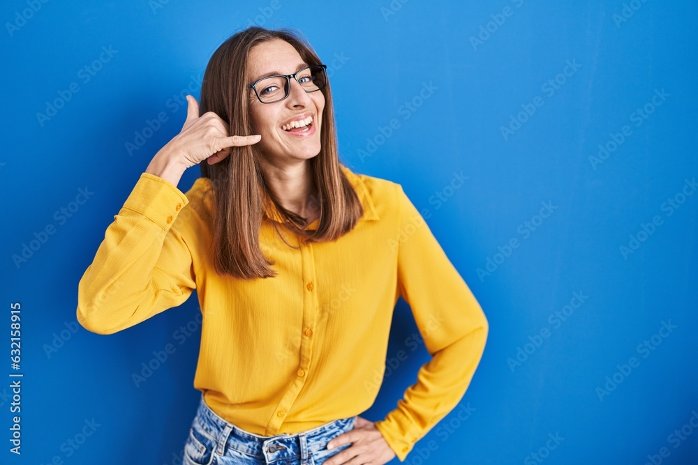 Young woman wearing glasses standing over blue background smiling doing phone gesture with hand and fingers like talking on the telephone. communicating concepts.