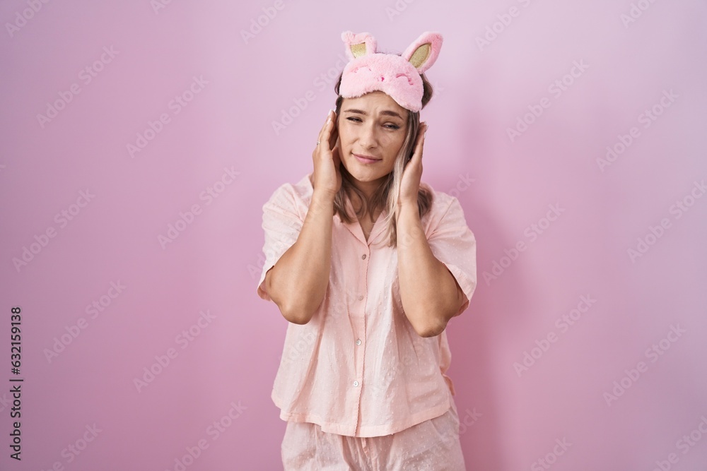 Blonde caucasian woman wearing sleep mask and pajama covering ears with fingers with annoyed expression for the noise of loud music. deaf concept.