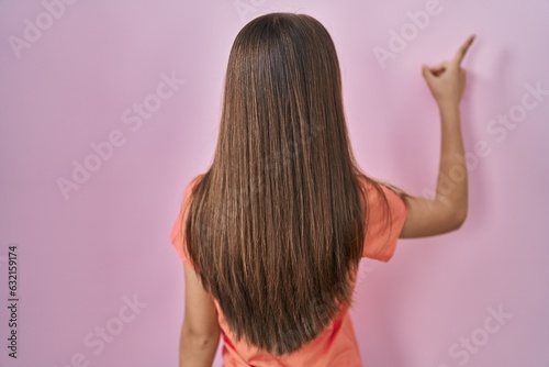 Teenager girl standing over pink background posing backwards pointing ahead with finger hand
