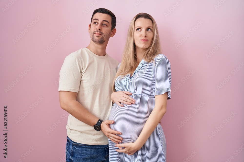 Young couple expecting a baby standing over pink background smiling looking to the side and staring away thinking. Young couple expecting a baby standing over pink background smiling looking to the side and staring away thinking.