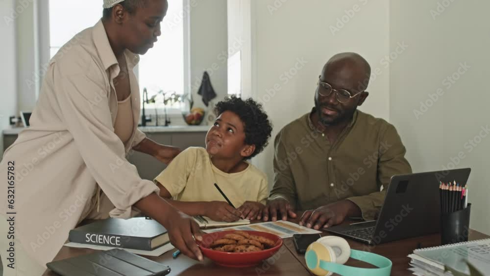 Waist up of African American father and preteen son studying together at home while mother serving homemade cookies at dining table on lunch time Waist up of African American father and preteen son studying together at home while mother serving homemade cookies at dining table on lunch time