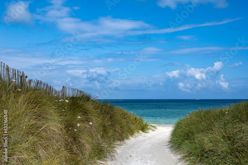 A dreamlike sandy path leads through green dunes to the turquoise sea in the north of Brittany under a blue summer sky.