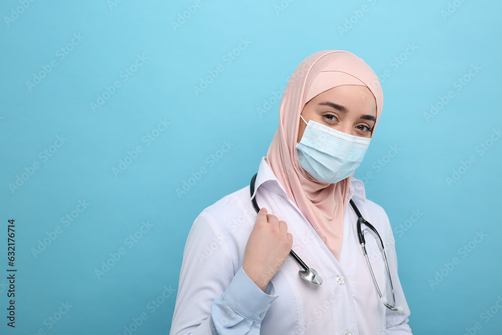 Muslim woman wearing hijab, medical uniform and protective mask on light blue background, space for text Muslim woman wearing hijab, medical uniform and protective mask on light blue background, space for text