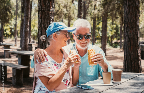 Smiling senior family couple enjoying a sandwich while sitting at a wooden table in the woods appreciating nature and freedom, happy retired seniors man and woman and healthy lifestyle concept