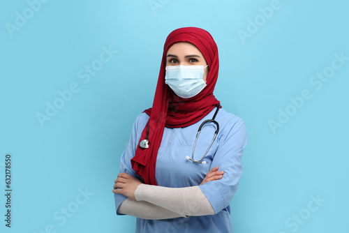 Muslim woman wearing hijab, medical uniform and protective mask on light blue background