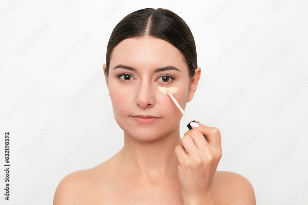 A young attractive Caucasian dark-haired woman applying a concealer under her eyes from dark circles under her eyes. Isolated on a light background A young attractive Caucasian dark-haired woman applying a concealer under her eyes from dark circles under her eyes. Isolated on a light background