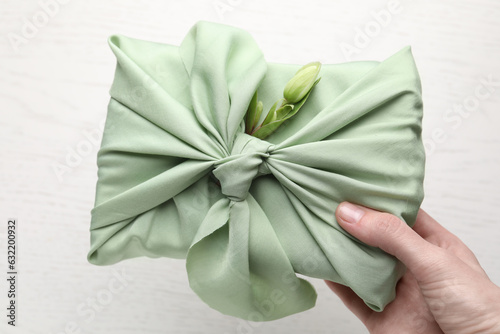 Furoshiki technique. Woman holding gift packed in green fabric with hellebore flower over light table, top view