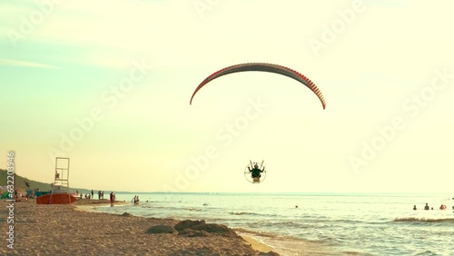 Man flying and gliding paraglider with motor above public sea beach at sunset. Resting active healthy lifestyle and wellness