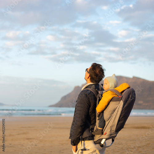 Father rising hands to the sky while enjoying pure nature carrying his infant baby boy son in backpack on windy sandy beach of Famara, Lanzarote island, Spain at sunset. Family travel concept © kasto