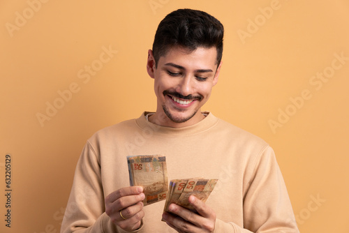 happy brazilian man looking at brazilian money cash in beige studio background. financial, credit, purchase, rich concept. 