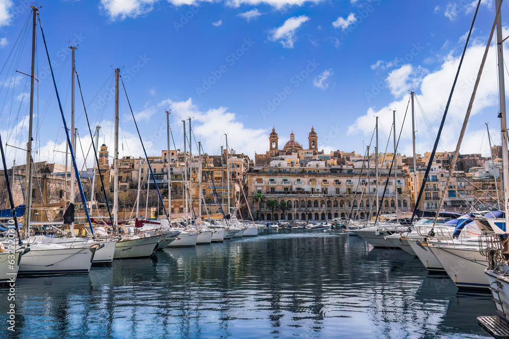Senglea, Malta waterfront with moored leisure boats and traditional limestone buildings with balconies against blue sky with clouds.