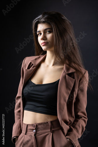 Classic studio portrait of a young brunette dressed in a black top and formal suit, who is sitting on a chair against a black background.