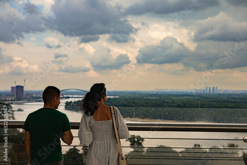 A man and a woman on a glass bridge look at the city. Rain and clouds. Back view