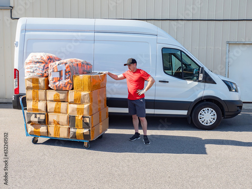 Man near cart with boxes. Delivery goods to factory. Loader near minivan. Guy loader is getting ready to load boxes into car. Man warehouse contractor. Male loader controls receipt goods at warehouse photo