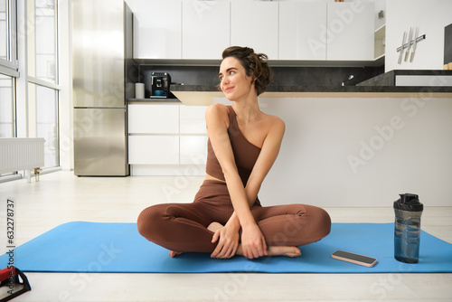 Portrait of fitness girl doing yoga on rubber mat at home, workout indoors in kitchen wearing sportswear, practice minfulness, sitting in lotus pose with water bottle next to her photo