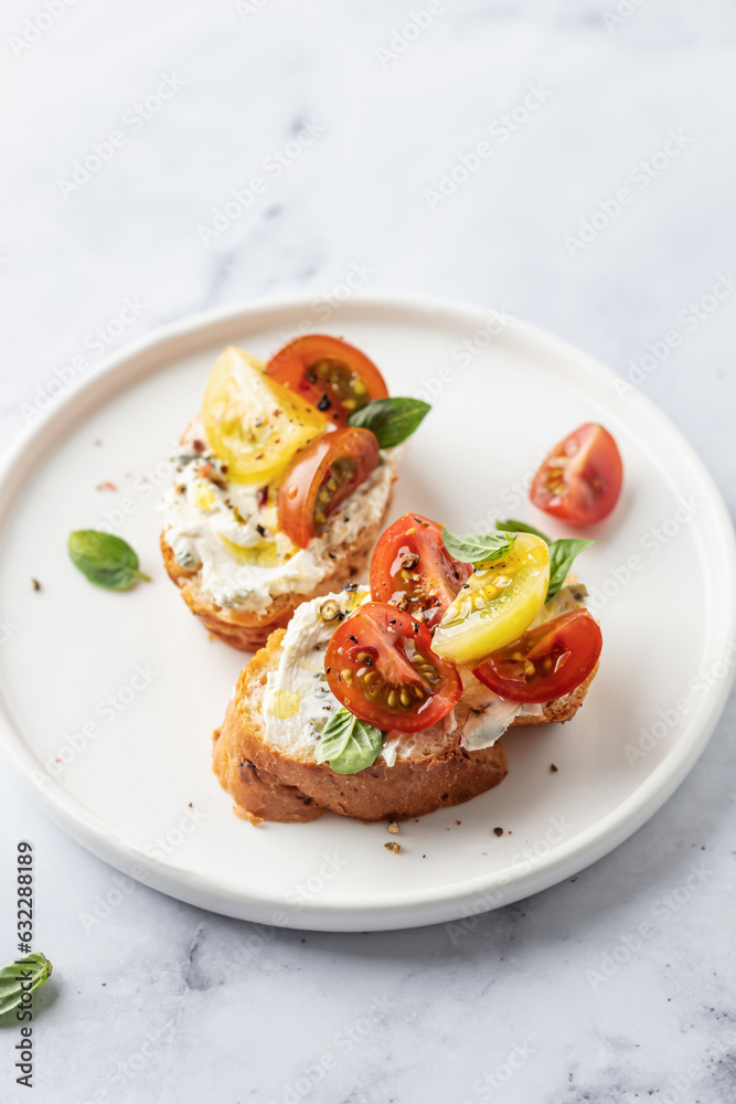 Bruschetta sandwiches with tomatoes, cream cheese, olive oil and basil on a plate on white marble background close up. Traditional italian antipasti