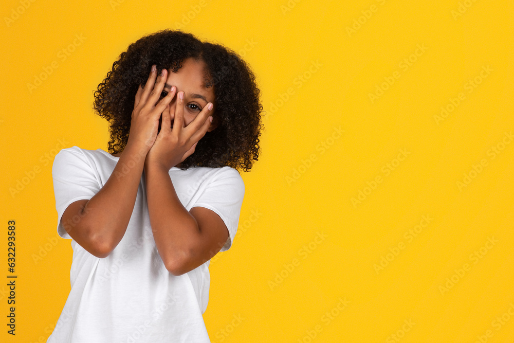 Shocked scared shy teenage black girl in white t-shirt covers face with hands, isolated on yellow background Shocked scared shy teenage black girl in white t-shirt covers face with hands, isolated on yellow background
