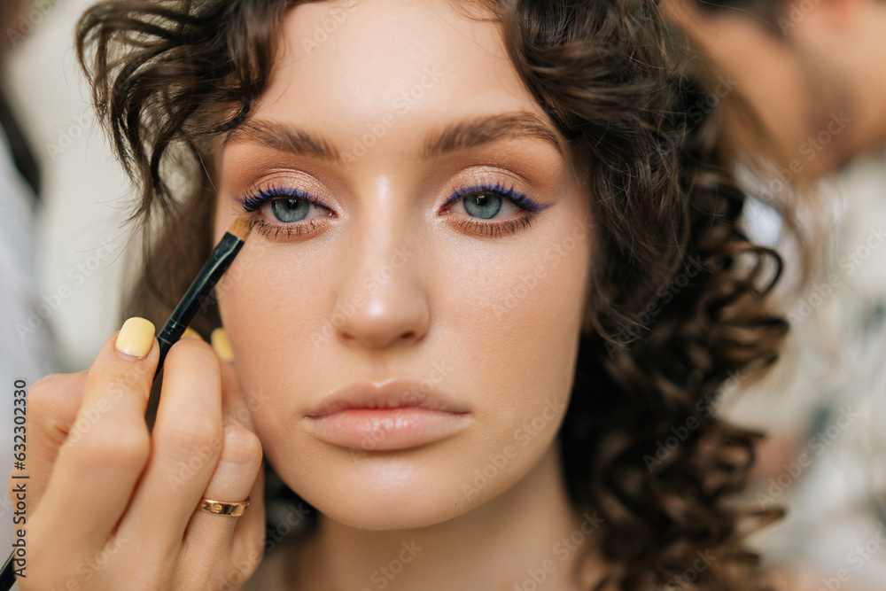 Close-up beautiful face of young woman getting makeup eyeliner with color shadows in modern beauty studio. Visagist using brushes facial professional cosmetics for makeup. Concept of beauty make-up.