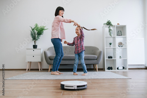 Εκτύπωση καμβά Joyful mother turning small kid around to energetic tunes while robot cleaner dealing with dirt on floor in apartment