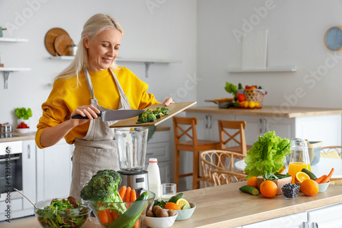 Mature woman with cut avocado making healthy smoothie in kitchen