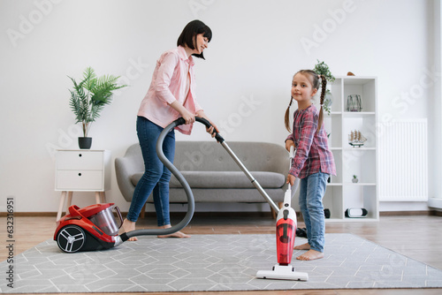 Full length view of preteen girl holding upright vacuum cleaner while female grown-up using canister model alongside. Prudent ladies of house collecting dust on various surfaces at sunny weekend.
