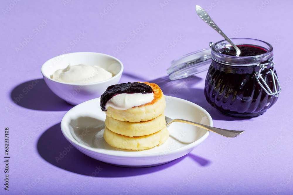 Breakfast of syrniki with sour cream and blackcurrant jam with orange on a lilac background.