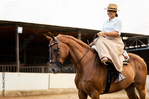 A cute girl in neat clothes is up on her brown horse with an amazon mount on the outside of the stable. The animal is of the Algo-Arabian breed.Young women riding horses. ©  Yistocking