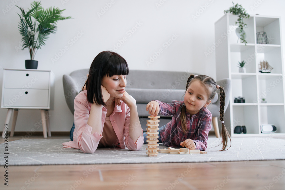 Attentive preteen girl putting block in tumble tower while calm adult lying alongside on carpet in apartment. Carefree parent and child taking turns moving components of toy building indoors. Attentive preteen girl putting block in tumble tower while calm adult lying alongside on carpet in apartment. Carefree parent and child taking turns moving components of toy building indoors.
