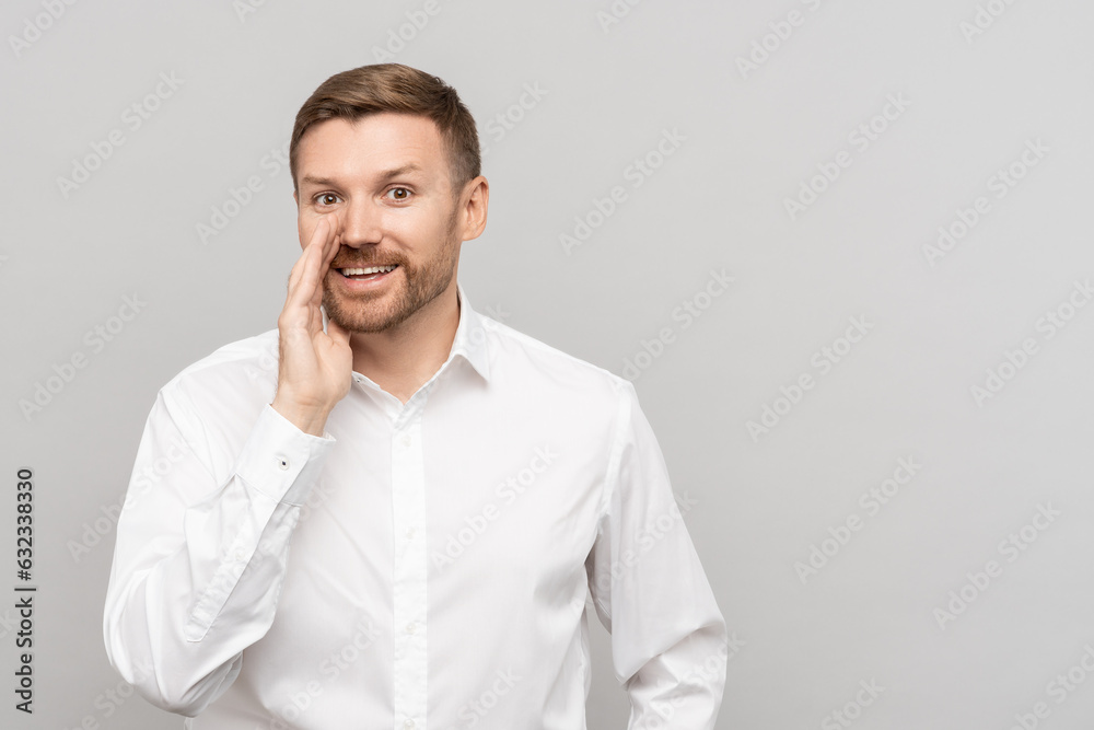 Positive man whispers promotional offer put palm to face on gray wall studio background. Pleased male standing at wall telling top secret reproduced for advertising in business white shirt copy space Positive man whispers promotional offer put palm to face on gray wall studio background. Pleased male standing at wall telling top secret reproduced for advertising in business white shirt copy space