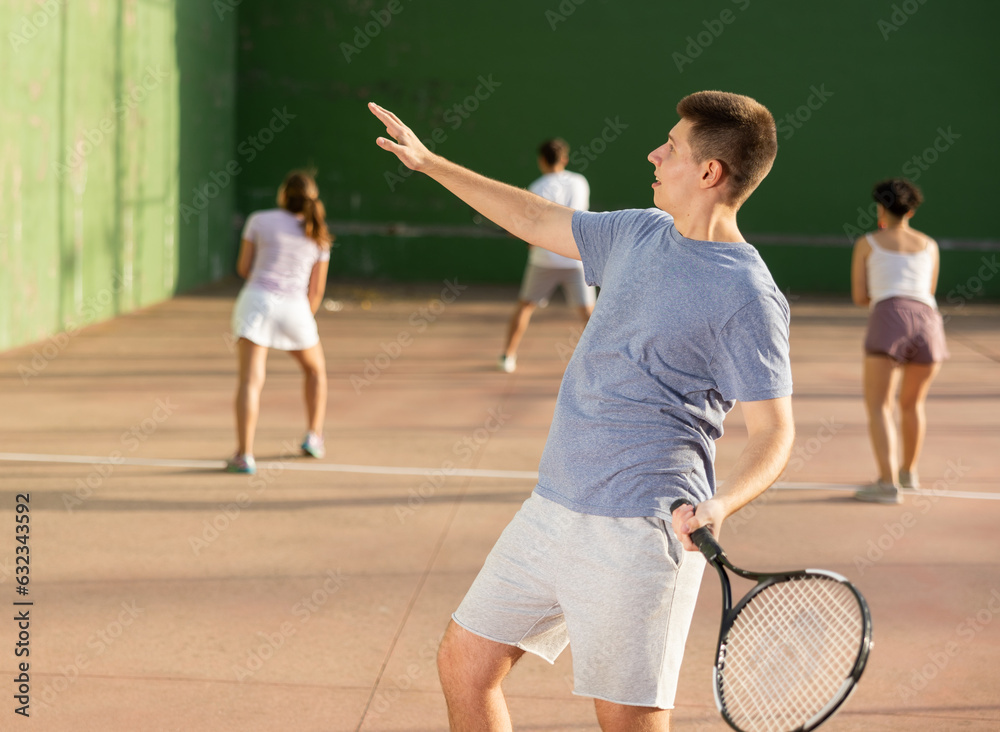 Sporty young man playing popular team game frontenis at open-air fronton court on summer day, ready to hit rubber ball with racquet